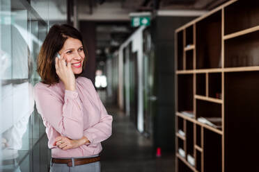 A portrait of businesswoman with smartphone standing in an office building with crossed arms, making a phone call. - HPIF18404