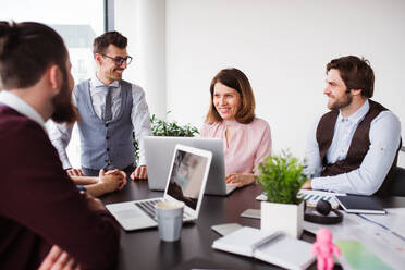 A group of young cheerful business people with laptop sitting in an office, talking. - HPIF18418