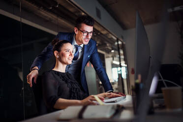 Two young business people in an office in the evening or at night, using computer. - HPIF18488