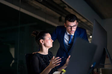 Two young business people in an office in the evening or at night, using computer. - HPIF18496