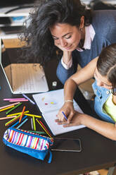 Freiberufliche Mutter, die ihrer Tochter beim Zeichnen in einem Notizbuch am Tisch hilft - JAQF01226