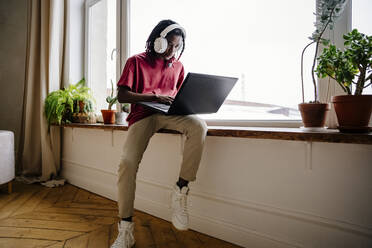 Junger Mann mit Laptop auf der Fensterbank zu Hause - NLAF00003