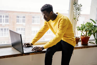 Konzentrierter Mann mit Laptop auf der Fensterbank zu Hause - NLAF00006