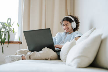 Glücklicher Junge mit Headset und Laptop auf der Couch zu Hause - NLAF00014