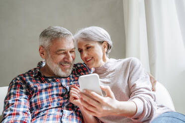 Smiling mature couple with smart phone sitting on couch at home - MDOF01118