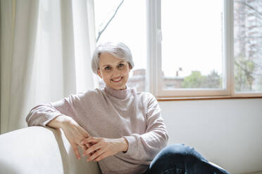 Smiling mature woman sitting on couch by window at home - MDOF01139