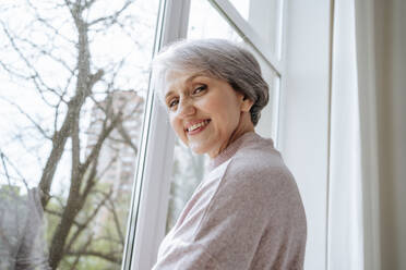 Reife Frau mit grauem Haar in der Nähe des Fensters zu Hause - MDOF01146