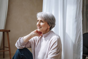 Thoughtful smiling woman sitting by curtain at home - MDOF01148