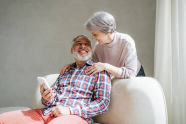 Älteres Paar, das sich gegenseitig anlächelt und ein Smartphone zu Hause in der Hand hält - MDOF01152