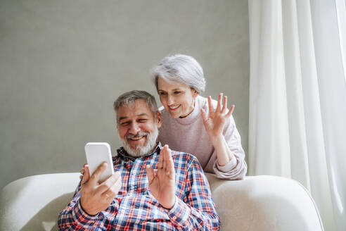 Lächelndes reifes Paar winkt bei einem Videoanruf über ein Smartphone zu Hause - MDOF01158