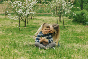 Glückliche Mutter umarmt und küsst Sohn sitzt auf Gras im Garten - VSNF00912