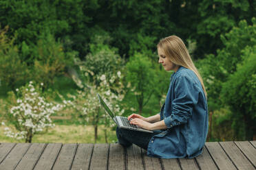Blond woman using laptop sitting on wooden boardwalk in garden - VSNF00916