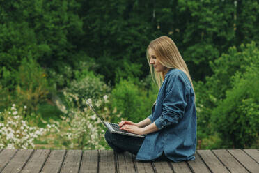 Woman using laptop sitting on wooden boardwalk in garden - VSNF00918