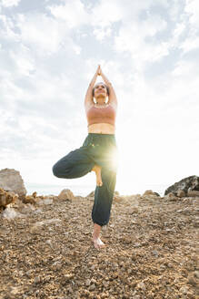 Frau übt Yoga in Baumhaltung vor dem Himmel am Strand - AAZF00535