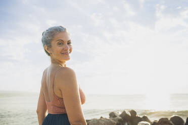 Lächelnde reife Frau schaut über die Schulter vor dem Himmel am Strand - AAZF00539