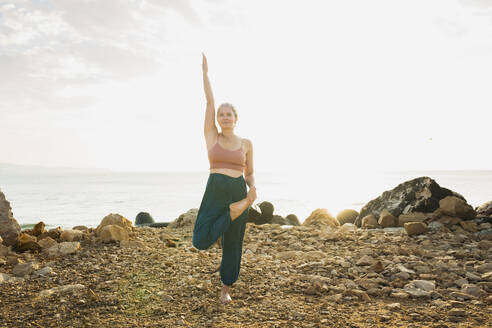 Frau übt Yoga auf einem Bein stehend vor dem Himmel am Strand - AAZF00540