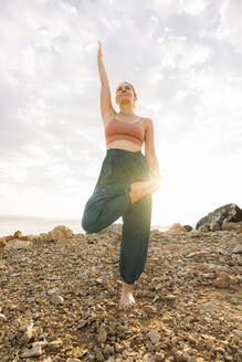 Frau übt Yoga auf einem Bein stehend am Strand - AAZF00541