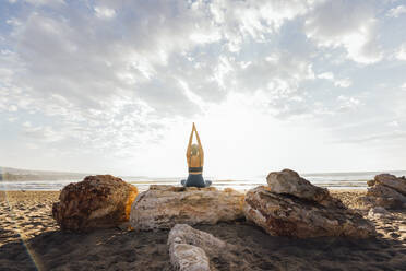 Woman practicing yoga sitting on rock at beach in front of sky - AAZF00545