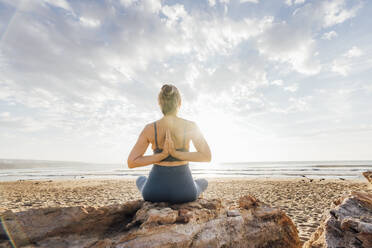 Woman practicing yoga with hands clasped behind back sitting on rock at beach - AAZF00546