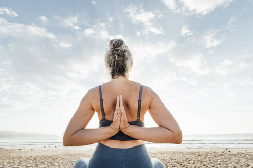 Woman practicing yoga with hands clasped behind back sitting at beach - AAZF00547