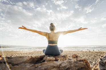 Woman practicing yoga with arms outstretched sitting on rock at beach in front of sky - AAZF00548