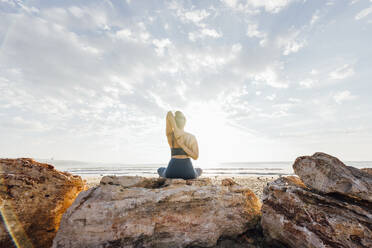 Frau übt Yoga mit den Händen hinter dem Rücken auf einem Felsen am Strand sitzend - AAZF00549