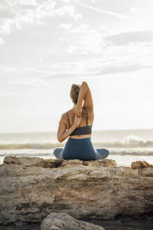 Woman doing yoga with hands behind back sitting on rock at beach - AAZF00550