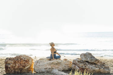 Frau übt Yoga auf einem Felsen am Strand vor dem Himmel sitzend - AAZF00554