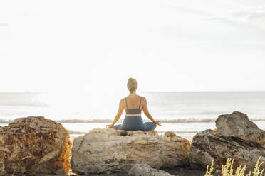 Woman meditating on rock at beach in front of sky - AAZF00555