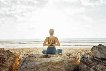 Woman practicing yoga with hands clasped behind back sitting at beach - AAZF00557