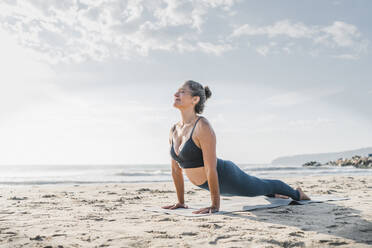 Woman practicing yoga with cobra pose in front of sky at beach - AAZF00561