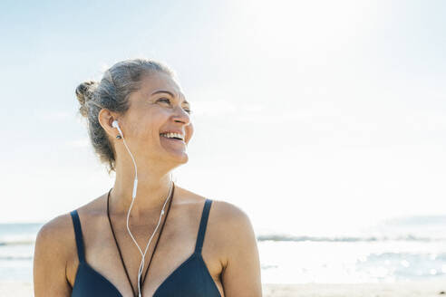 Glückliche Frau mit In-Ear-Kopfhörern, die am Strand nachdenkt - AAZF00580