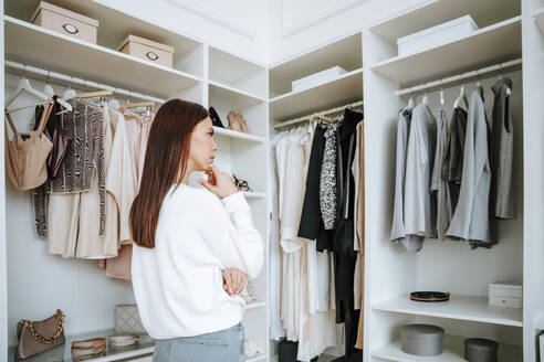 Thoughtful woman with hand on hip looking at clothes hanging in wardrobe - NLAF00023