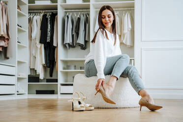 Smiling woman trying heels sitting on seat in closet - NLAF00029