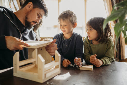 Sons looking at father building up birdhouse at home - ANAF01442