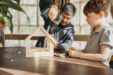 Father drilling birdhouse on table with son at home - ANAF01447
