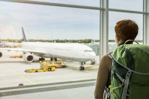 Frau mit Rucksack betrachtet Flugzeug durch Fenster am Flughafen - MAMF02860