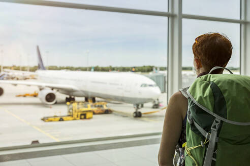 Woman with backpack looking at plane through window at airport - MAMF02860