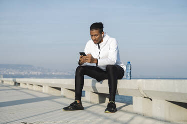 Athlete using smart phone sitting on promenade bench - ALKF00305