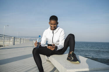 Lächelnder Sportler mit Smartphone auf einer Bank am Meer sitzend - ALKF00306
