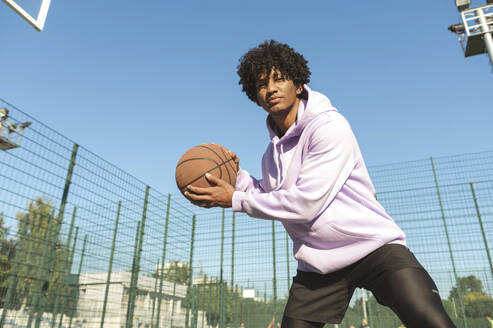 Sportler mit lockigem Haar, der einen Basketball auf einem Sportplatz hält - ALKF00316