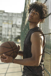 Junger Sportler hält Basketball an einen Zaun gelehnt - ALKF00319