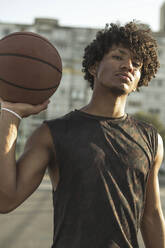 Selbstbewusster Sportler mit lockigem Haar, der einen Basketball auf einem Sportplatz hält - ALKF00322