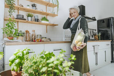 Smiling woman looking at mesh bag with groceries standing in kitchen at home - YTF00829