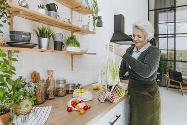 Lächelnde Frau, die eine Netztasche voller Lebensmittel auf der Theke in der Küche zu Hause aufbewahrt - YTF00830