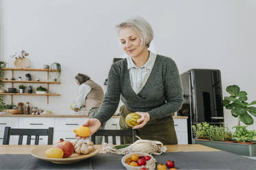 Smiling woman arranging fruits on dining table in kitchen at home - YTF00834