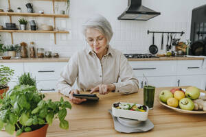Ältere Frau mit Tablet-PC in der Küche zu Hause sitzend - YTF00878