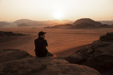 Junger Mann mit Mütze sitzt auf einem Felsen in der Wüste bei Sonnenuntergang - PCLF00569