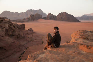 Happy young man sitting on rock in the desert at sunset - PCLF00570