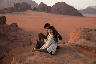 Smiling woman and man sitting with cat on rock in the desert at sunset - PCLF00571